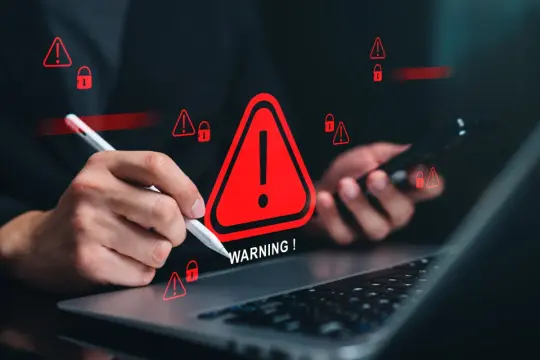 Ręce nad laptopem i telefonem, otoczone czerwonymi ostrzeżeniami i ikonami kłódki. To jakby tracker kto to, próbuje się włamać.