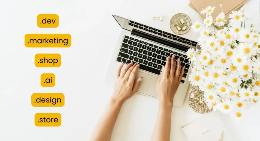 Kobieta pisze na laptopie, obok bukiet margaretek. Widoczne są różne rozszerzenia domen, np. .dev, .marketing, .shop, .ai, .design, .store.