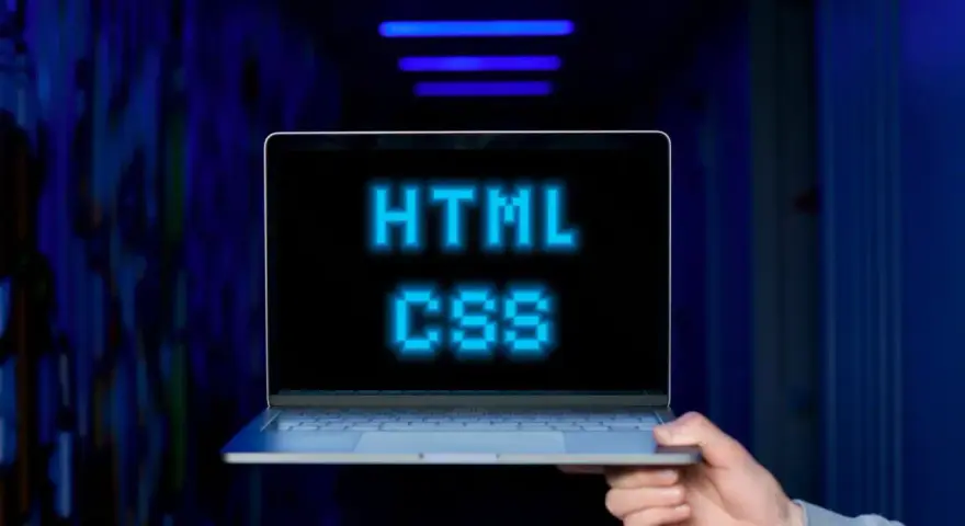 <p>Dłoń trzyma laptopa, na ekranie którego świeci napis "HTML CSS" w niebieskich pikselach. Tło jest ciemne, z niebieskimi neonami.</p>